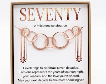 May include: A rose gold necklace with seven interlocking circular rings displayed on a white card inside a brown box. The card features the text SEVENTY, A Milestone celebration, and a message about seven rings representing seven decades.