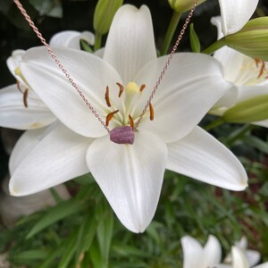 May include: A delicate rose gold chain necklace with a small, dark purple gemstone pendant. The necklace is displayed against a backdrop of white lilies.