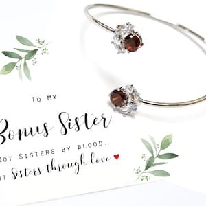 May include: A silver bangle bracelet with two red gemstone accents. The bracelet is accompanied by a card that reads "To my Bonus Sister, Not sisters by blood, but sisters through love."