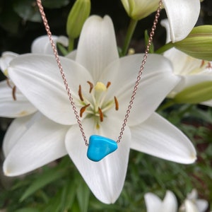 May include: A delicate rose gold chain necklace with a turquoise stone pendant. The necklace is photographed against a backdrop of white lilies.