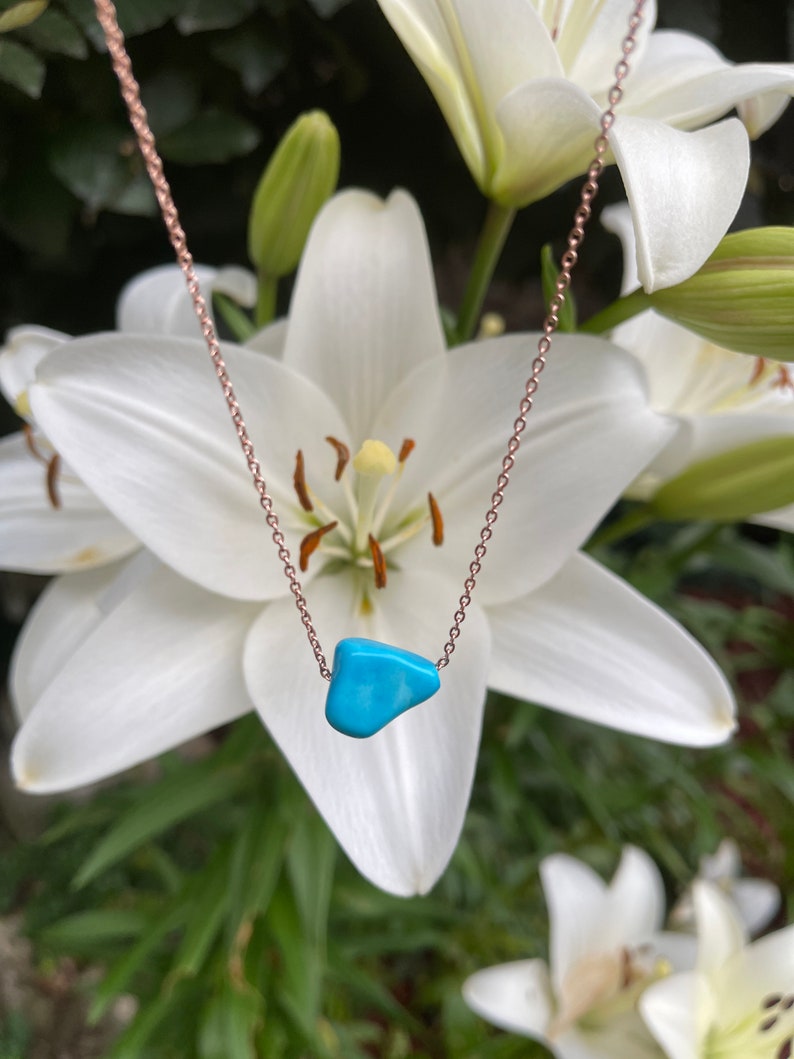 May include: A delicate rose gold chain necklace with a turquoise stone pendant. The necklace is photographed against a backdrop of white lilies.