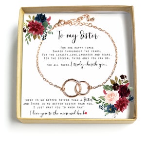 May include: A gift box with a rose gold bracelet featuring two interlocking circles. The box has a floral design with burgundy, pink, and blue flowers. The text on the card reads "To my Sister" and includes a message about cherishing the bond between sisters.