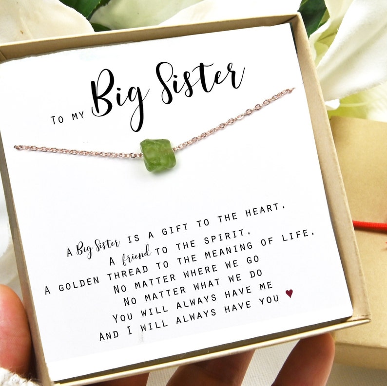 May include: A white card with the text "To My Big Sister" and a quote about the importance of a big sister. A delicate gold chain with a green gemstone pendant is displayed on the card.