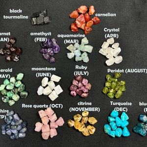 May include: A collection of small, rough gemstones arranged in rows, labelled with their corresponding birth month and name. The gemstones include black tourmaline, carnelian, garnet, amethyst, aquamarine, crystal, emerald, moonstone, ruby, peridot, sapphire, rose quartz, citrine, turquoise, and blue stone.