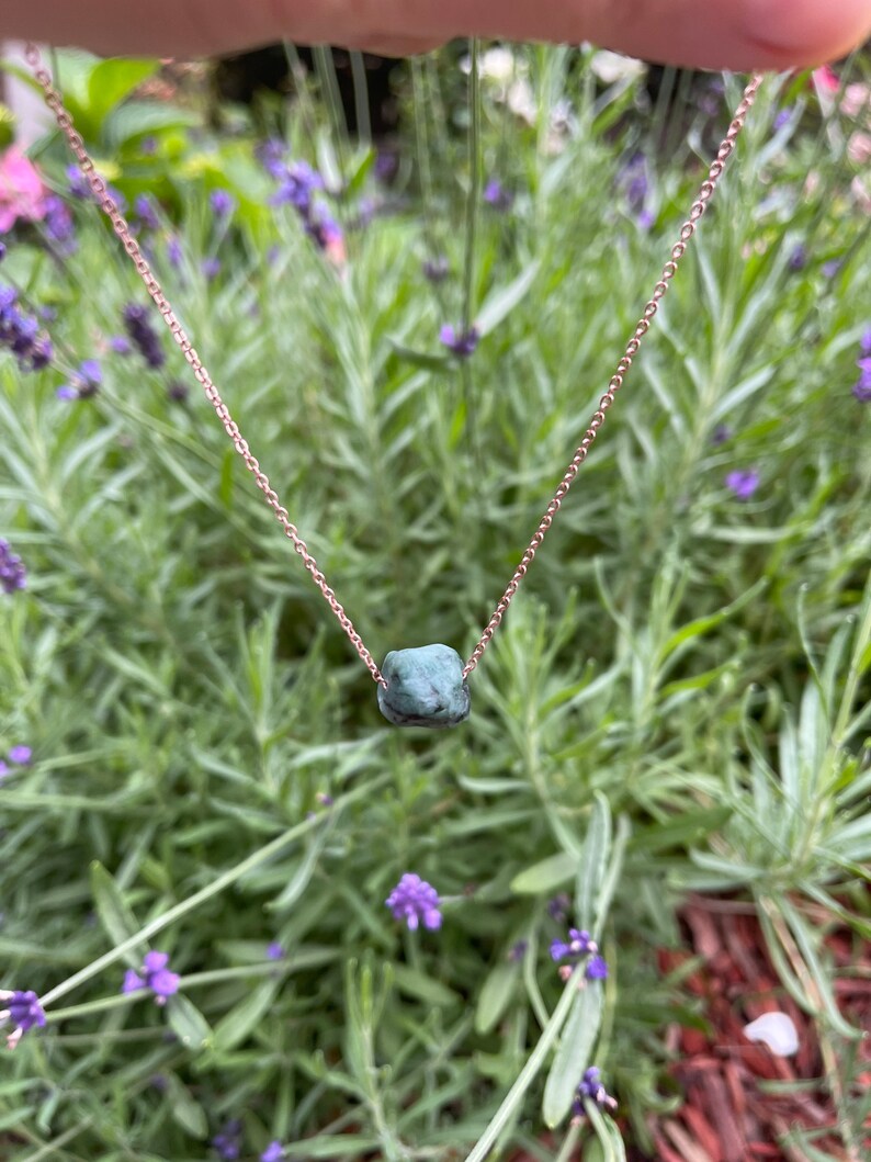 May include: A delicate gold chain necklace with a single, dark green, rough-cut gemstone pendant.