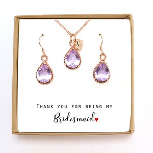 May include: A gift box with a rose gold necklace and earrings featuring a teardrop-shaped purple gemstone. The box has a card that reads "Thank you for being my Bridesmaid".