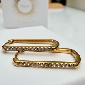 May include: Gold-toned rectangular hoop earrings with a row of small, clear crystals along the bottom edge. The earrings are displayed on a white surface, with a blurred white box in the background.