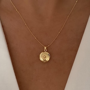 May include: A delicate gold necklace featuring a round pendant with a small, centered design. The chain is adorned with tiny beads, and the pendant hangs gracefully. The necklace is displayed against a white background.