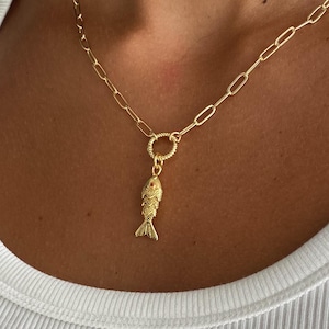 May include: Gold-colored necklace featuring a fish pendant. The necklace has a delicate chain with a small fish charm. The fish pendant has detailed scales and fins. The necklace is worn on a person.