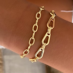 May include: Two gold-colored chain bracelets are displayed on a person's arm. One bracelet features oval links, while the other has elongated, open links. The bracelets are made of a shiny, yellow metal.