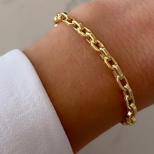 May include: A gold-colored chain link bracelet. The bracelet is made of a series of rectangular links. The bracelet is worn on a wrist, with a white shirt sleeve visible in the background. The Jewelry Standard logo is in the bottom right corner.