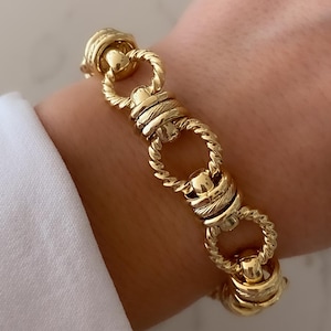 May include: Gold-toned bracelet featuring a series of interconnected, textured links. The bracelet has a polished finish, with a combination of twisted rope and smooth, rounded elements. The design is visible against a white shirt sleeve and a light background.