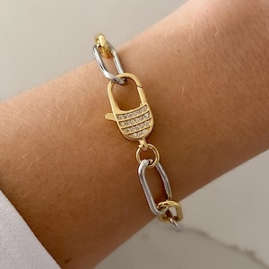 May include: A gold and silver chain bracelet with a decorative clasp. The clasp is gold and encrusted with small, clear stones. The bracelet is worn on a wrist, and the background is a light, neutral color.