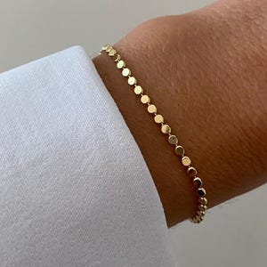 May include: A delicate gold bracelet composed of a continuous chain of small, flat, circular metallic discs. The bracelet features a polished finish that reflects light, creating a uniform, linked design that encircles the wrist in a simple, linear fashion.