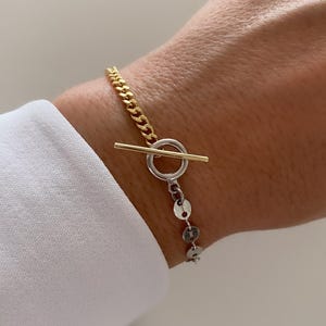 May include: A gold and silver bracelet with a toggle clasp. The bracelet features a gold chain and a silver chain with small, round, disc-shaped charms. The clasp is a gold bar that fits through a silver ring.