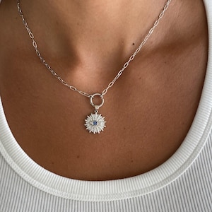 May include: A silver necklace with a pendant shaped like a sunburst. The pendant has a blue center stone and is surrounded by smaller, clear stones. The necklace is worn on a person wearing a white ribbed tank top.