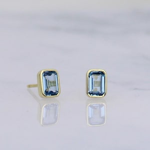 May include: A pair of stud earrings featuring rectangular, light blue gemstones set in gold-colored bezels. The earrings are displayed on a reflective surface, showcasing their shape and color. The gemstones have a subtle, transparent quality.