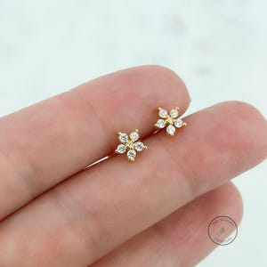 May include: A pair of gold-colored flower stud earrings. Each earring features a flower design with six clear, round-cut gemstones. The earrings are displayed on a person's finger.