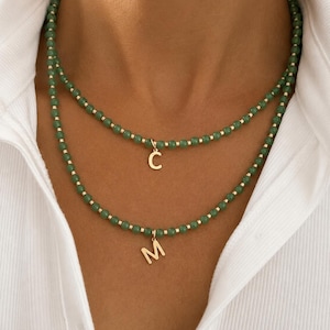 May include: A layered necklace set featuring two strands of small, round, green beads interspersed with tiny gold-toned cylindrical spacers. The shorter strand features a gold-toned letter C charm, while the longer strand features a gold-toned letter M charm.