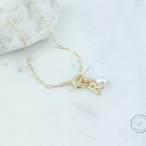 May include: A gold-colored chain necklace with a toggle clasp and a pendant. The pendant features the letter "R", a white pearl, and a small gold bead. The necklace is displayed on a white marble surface.