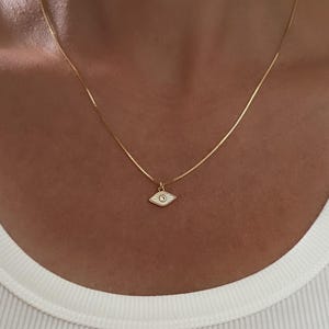 May include: A gold-colored necklace with a small, white and gold evil eye pendant. The pendant has a blue center and is suspended from a delicate chain. The necklace is worn against a white shirt.
