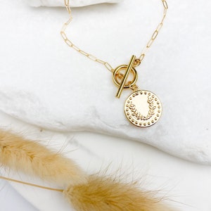 K&ouml;nnte beinhalten: Goldkette mit einem Schiebeverschluss und einem runden Anh&auml;nger mit einem floralen Design.