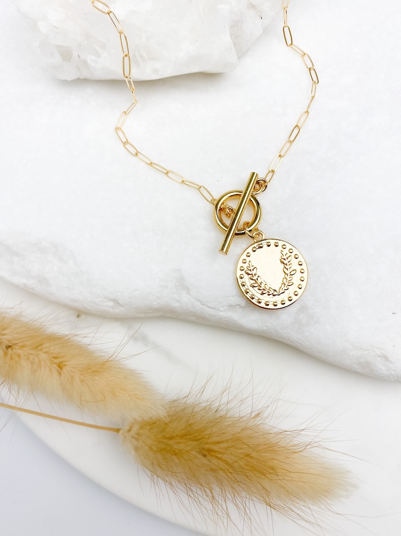 K&ouml;nnte beinhalten: Goldkette mit einem Schiebeverschluss und einem runden Anh&auml;nger mit einem floralen Design.