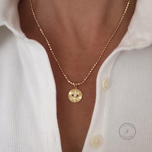 May include: Gold-toned necklace featuring a circular pendant with an eye design and a small, round, white gem in the center. The pendant is suspended from a delicate chain. The necklace is worn over a white collared shirt.