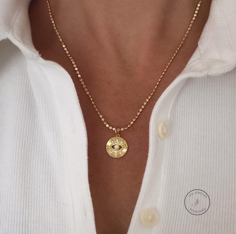 May include: Gold-toned necklace featuring a circular pendant with an eye design and a small, round, white gem in the center. The pendant is suspended from a delicate chain. The necklace is worn over a white collared shirt.