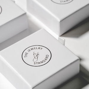 May include: White square boxes with a black circular logo that reads "The Jewelry Standard" with a stylized letter "J" inside the circle.