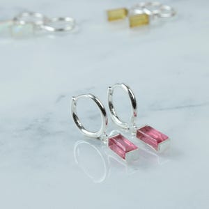 May include: A pair of silver hoop earrings with rectangular pink gemstone drops. The earrings are displayed on a white marble surface, with a blurred background showing additional jewelry.