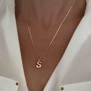 May include: A gold-colored necklace with a pendant in the shape of the letter "S". The pendant is a rounded, bubble-style font. The necklace is worn against a white shirt.