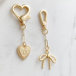 May include: Two gold-colored keychains. One keychain has a heart-shaped clasp and a heart pendant with the letter "D". The other keychain has a clasp and a bow pendant. Both keychains have a gold chain.