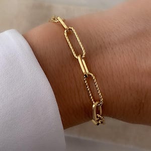 May include: A gold-colored link bracelet featuring a mix of oval and textured chain links. The bracelet is shown on a wrist, with a white shirt sleeve in the background. The bracelet's clasp is visible.