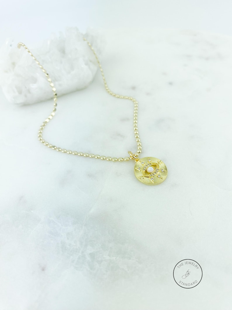 May include: Gold-toned necklace with a pendant featuring an eye design. The pendant is adorned with small, clear gemstones and a central pearl-like bead. The necklace is displayed on a white marble surface.