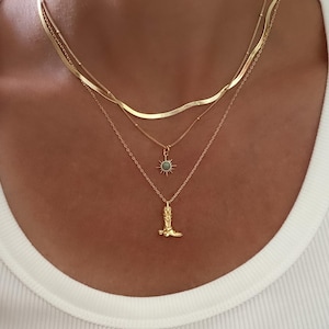 May include: A layered gold necklace set. The top layer is a flat, wavy chain, followed by a delicate beaded chain, and a chain with a sunburst pendant. The bottom layer features a gold cowboy boot pendant.