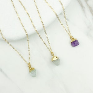 May include: Three gold chain necklaces with different colored crystal pendants. The necklaces are on a white marble surface. The crystals are a light green, a light blue, and a purple.