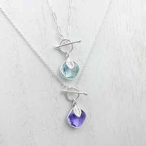 May include: Two silver necklaces with toggle clasps and square-shaped gemstone pendants. The top necklace has a light blue gemstone and the bottom necklace has a purple gemstone.