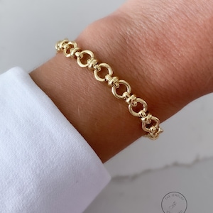 May include: A gold chain bracelet featuring a series of repeating circular links connected by small horizontal bars. The bracelet has a polished, reflective finish and is worn around the wrist. A small circular logo with the text THE JEWELRY STANDARD is visible in the bottom right corner.