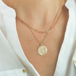 May include: A gold chain necklace with a large gold coin pendant featuring a detailed design. The necklace is layered over a thinner gold chain.