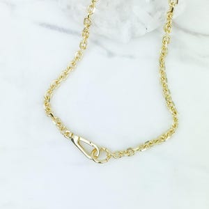 May include: A gold-colored chain necklace with a lobster clasp. The chain is made of oval links and is displayed on a white marble surface. The necklace is a simple, elegant piece of jewelry.