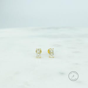 May include: A pair of gold stud earrings with rectangular, clear gemstones. The earrings are set on a white marble surface. The Jewelry Standard logo is visible in the lower right corner. These earrings are a classic jewelry item.