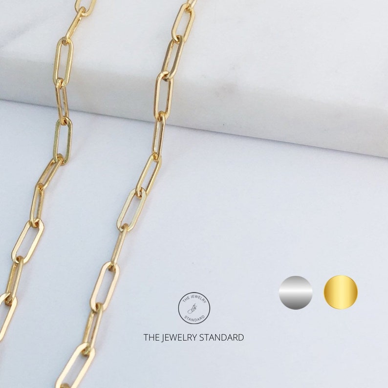 May include: A gold chain necklace with a delicate, rectangular link design. The chain is shown on a white marble surface.