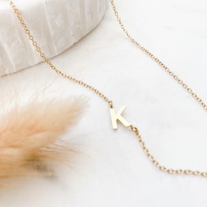 May include: A delicate gold-toned necklace featuring a single capital letter K pendant integrated directly into the fine link chain. The metal has a polished finish, and the letter is positioned asymmetrically along the length of the chain.