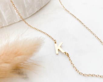May include: A delicate gold-toned necklace featuring a single capital letter K pendant integrated directly into the fine link chain. The metal has a polished finish, and the letter is positioned asymmetrically along the length of the chain.