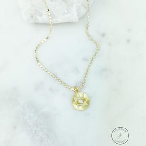 May include: Gold-colored necklace with a circular pendant featuring an eye design. The pendant has a central pearl and is surrounded by small, sparkling accents. The necklace chain is delicate and has a textured appearance. The Jewelry Standard logo is visible.