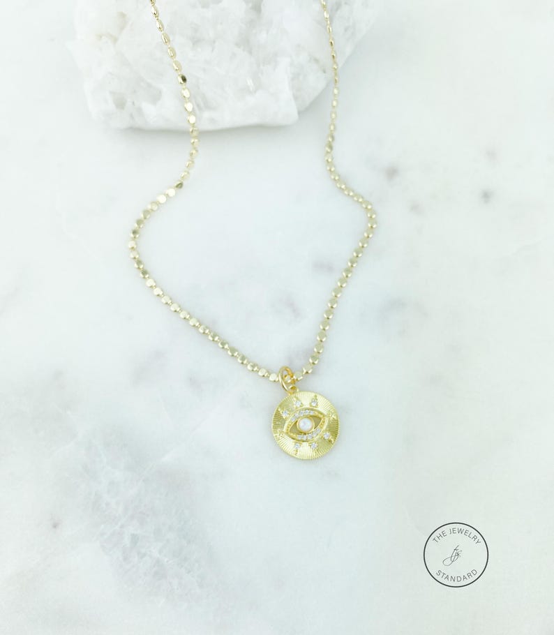 May include: Gold-colored necklace with a circular pendant featuring an eye design. The pendant has a central pearl and is surrounded by small, sparkling accents. The necklace chain is delicate and has a textured appearance. The Jewelry Standard logo is visible.