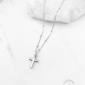 May include: A silver-colored necklace with a small cross pendant. The chain features small, round beads and a delicate design. The cross is centered and hangs from the chain. The Jewelry Standard logo is in the bottom right corner.