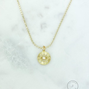 May include: Gold-toned necklace featuring a circular pendant with an eye design. The pendant has a central pearl and is surrounded by small, sparkling accents. The chain is a delicate, beaded style. The jewelry is displayed on a marble surface.