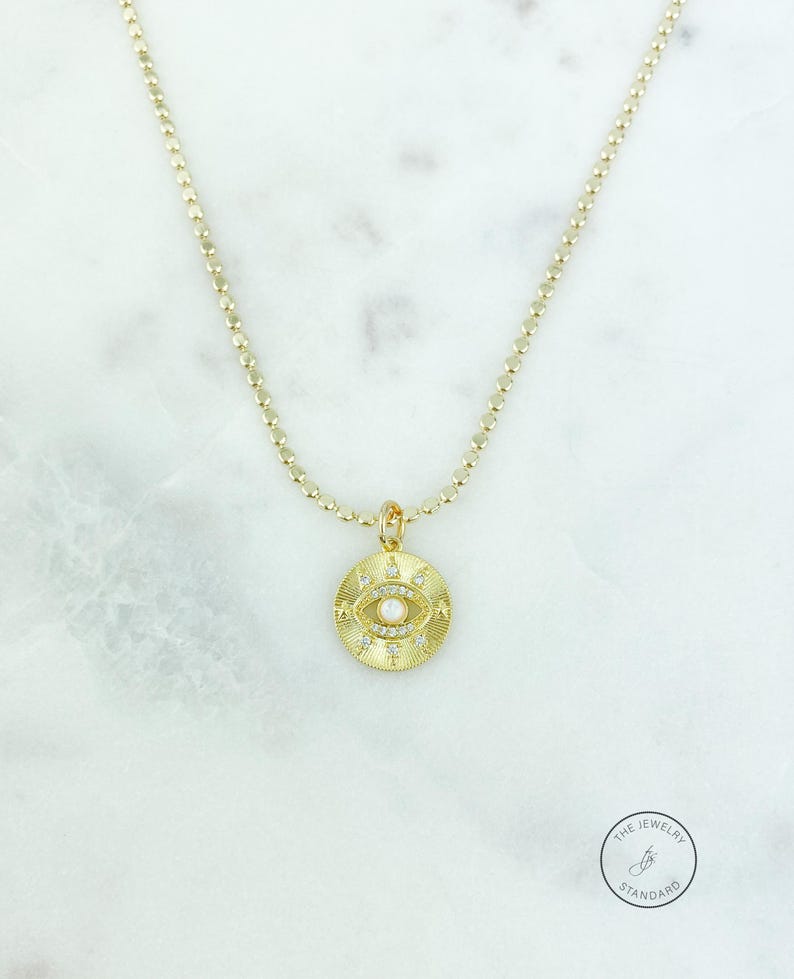 May include: Gold-toned necklace featuring a circular pendant with an eye design. The pendant has a central pearl and is surrounded by small, sparkling accents. The chain is a delicate, beaded style. The jewelry is displayed on a marble surface.